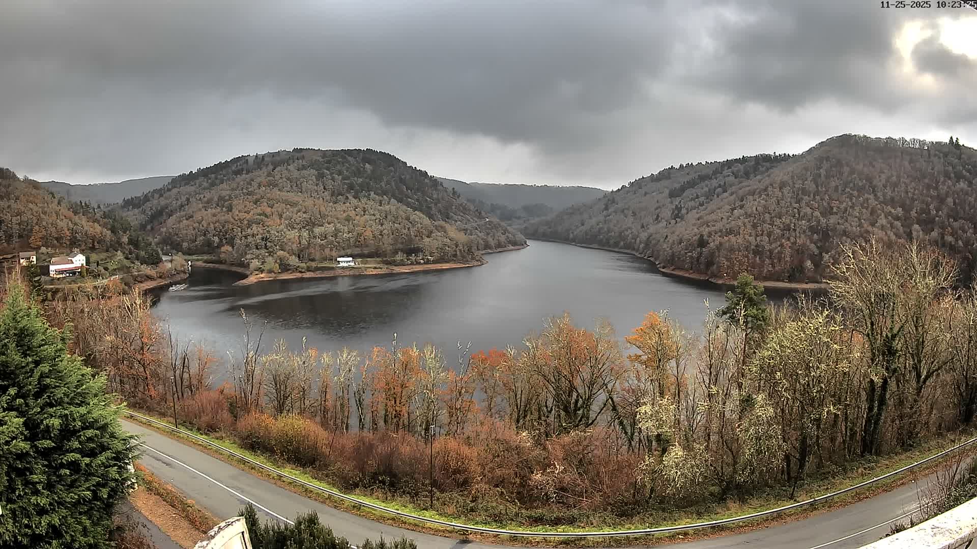 A winding, dark body of water is nestled between steep, tree-covered hills with late autumn foliage and a few scattered houses, all under a heavily overcast and gloomy sky.