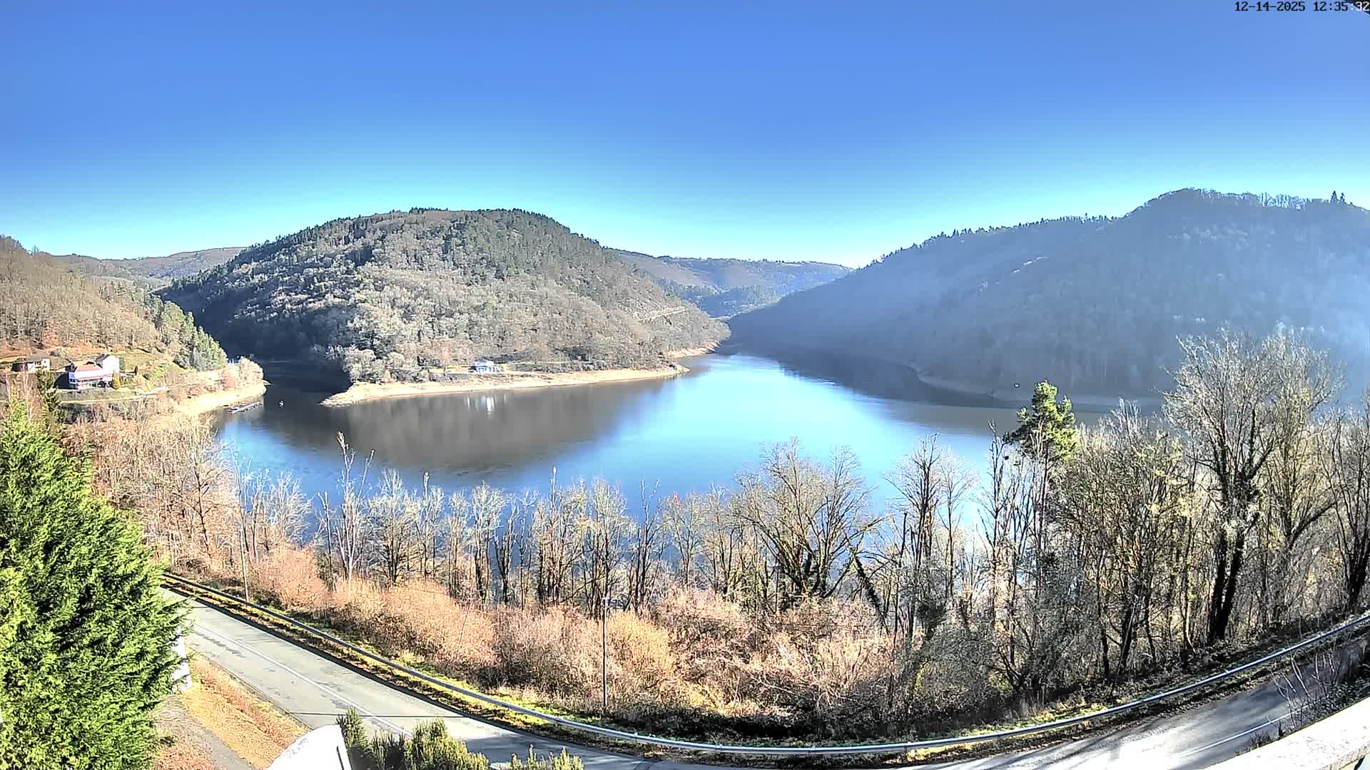 A winding, dark body of water is nestled between steep, tree-covered hills with late autumn foliage and a few scattered houses, all under a heavily overcast and gloomy sky.