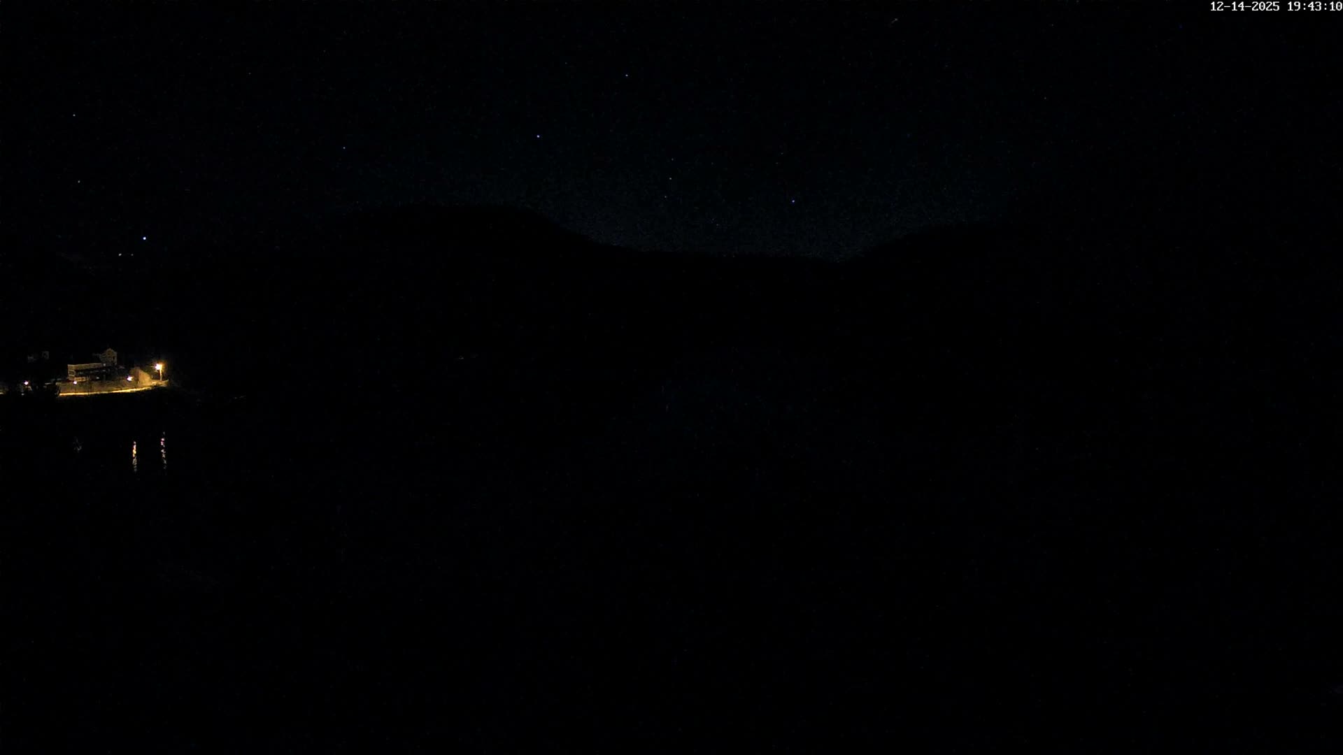 A winding, dark body of water is nestled between steep, tree-covered hills with late autumn foliage and a few scattered houses, all under a heavily overcast and gloomy sky.