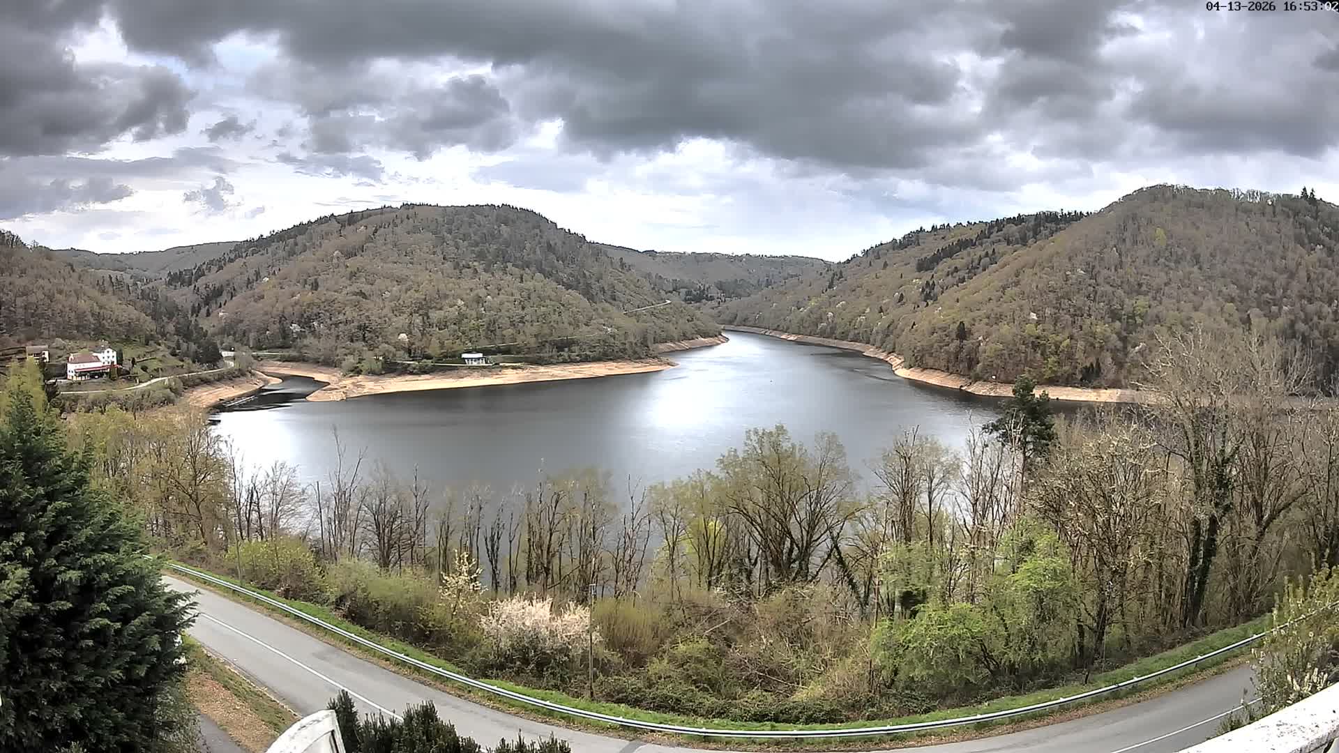 A winding, dark body of water is nestled between steep, tree-covered hills with late autumn foliage and a few scattered houses, all under a heavily overcast and gloomy sky.