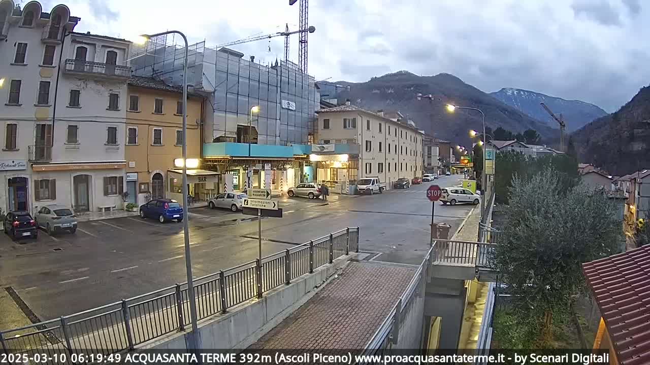 A town square with several parked cars, buildings under construction, and overcast skies in a mountainous region.