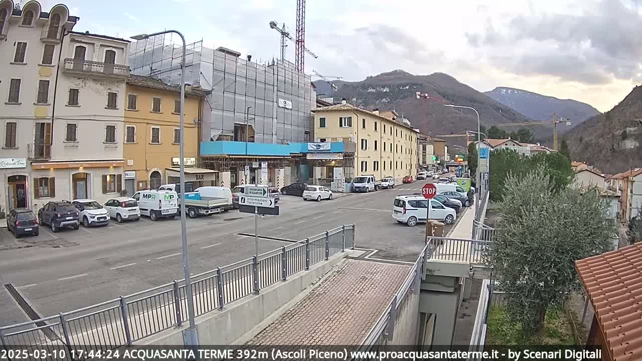 A street scene in a town nestled in a valley shows overcast skies, parked cars, buildings under construction, and mountains in the background.