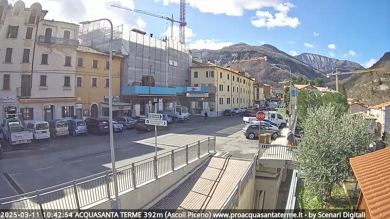 A sunny day reveals a town square with parked cars, buildings under construction, and mountains in the background.
