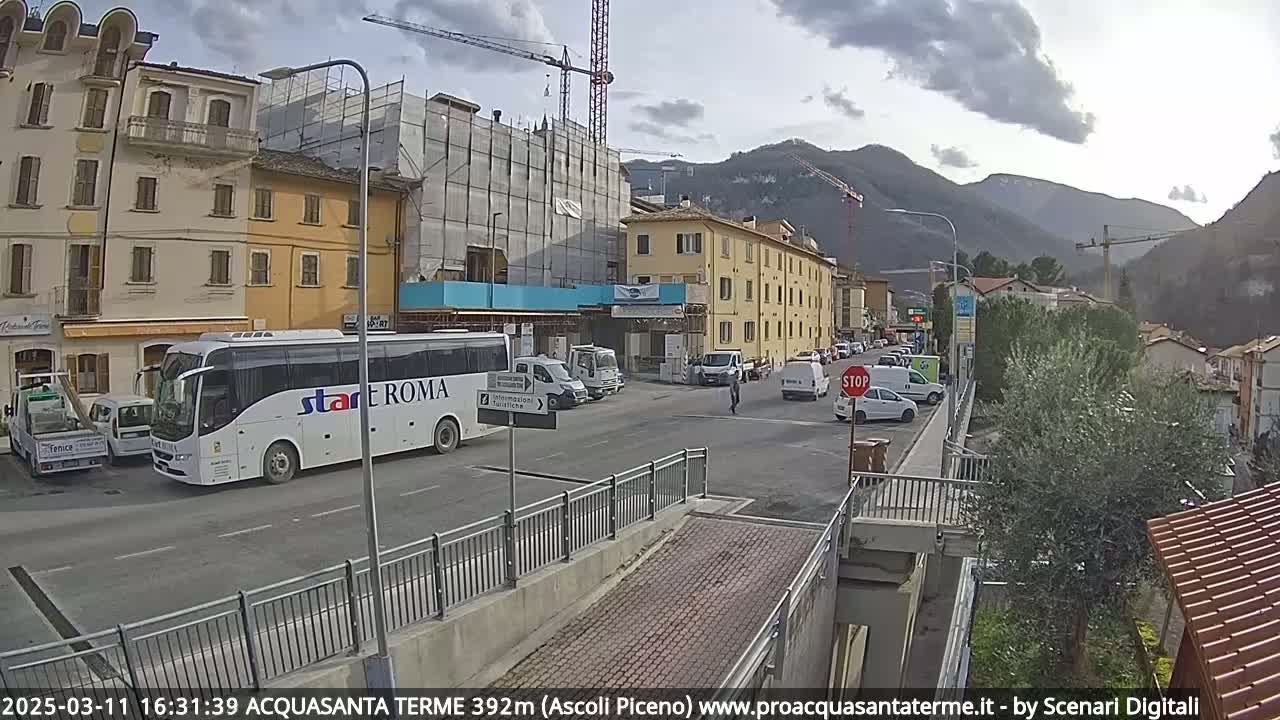 A street scene shows a large tour bus parked near several other vehicles, a construction site, and buildings under a partly cloudy sky with mountains in the background.