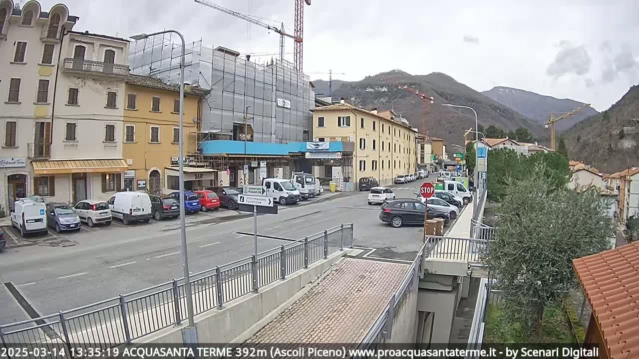 A street scene in a town nestled in a valley shows overcast weather, parked cars, buildings under construction, and a few cars driving on the road.