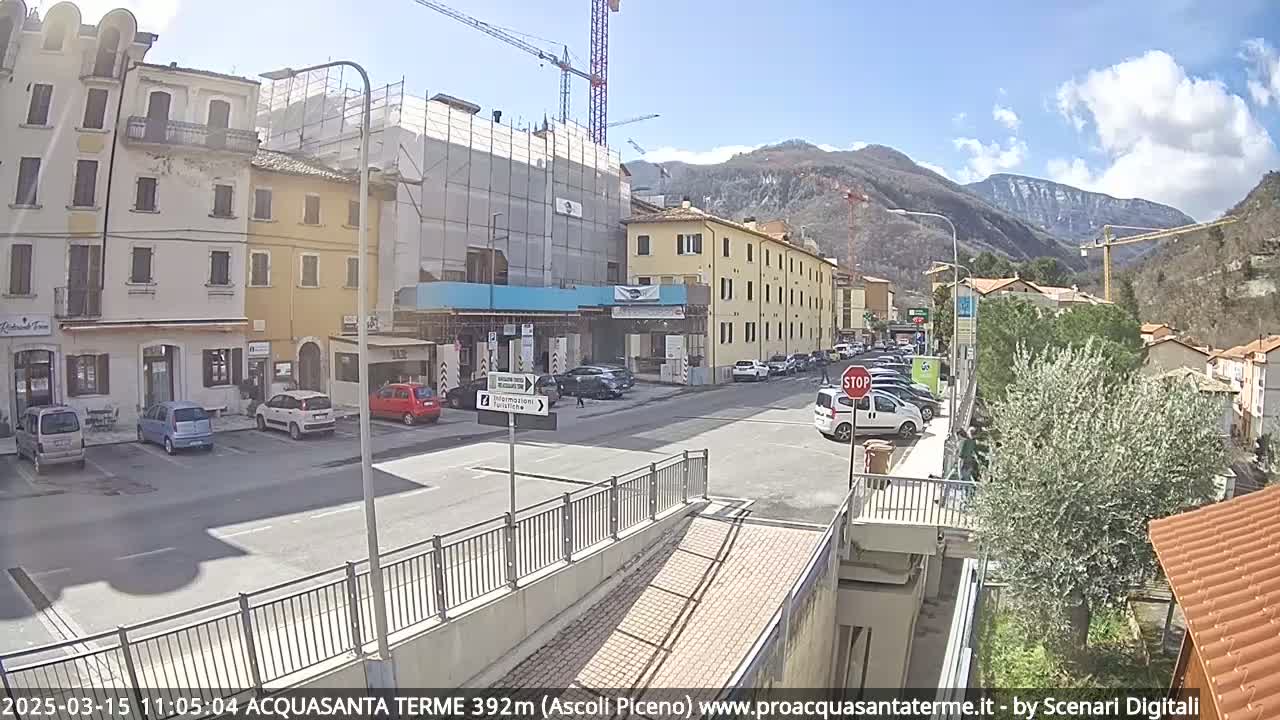 A sunny day reveals a street scene in a town nestled against mountains, with cars parked along the sides of buildings under construction.