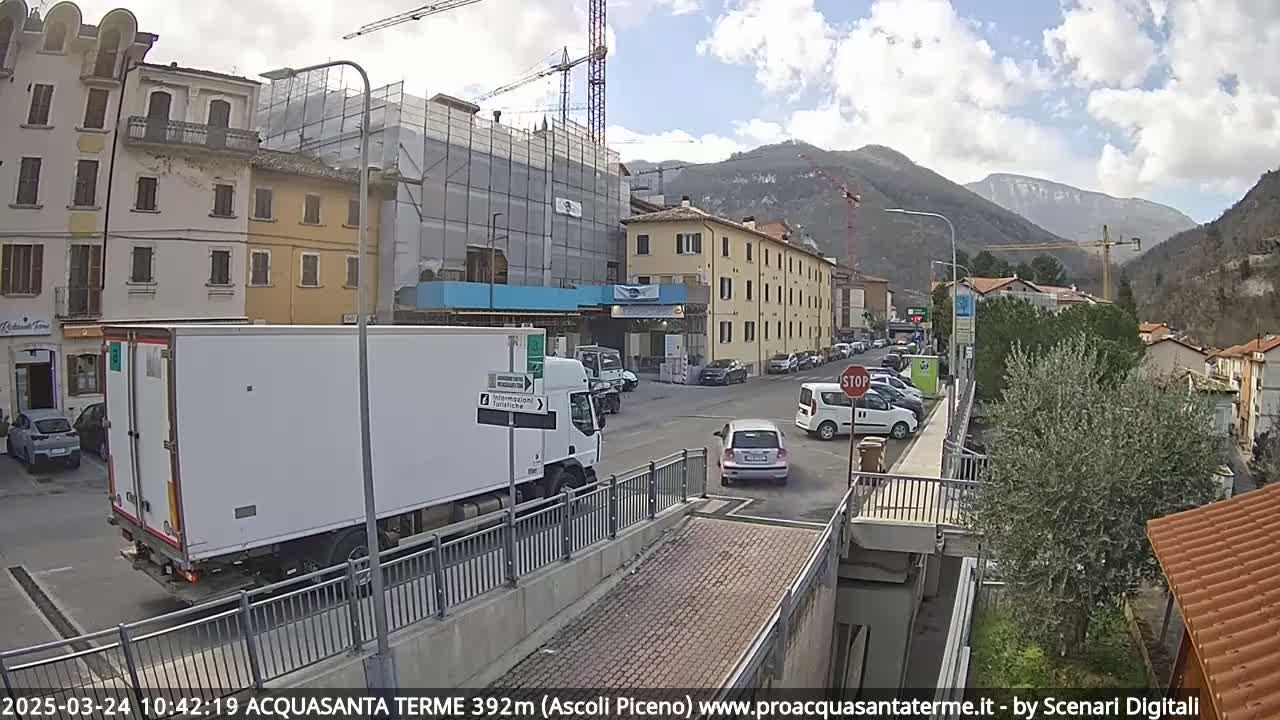 A partly cloudy day reveals a street scene in a town nestled in a valley between mountains, with a large white delivery truck and several cars parked along the roadside near a building under construction.