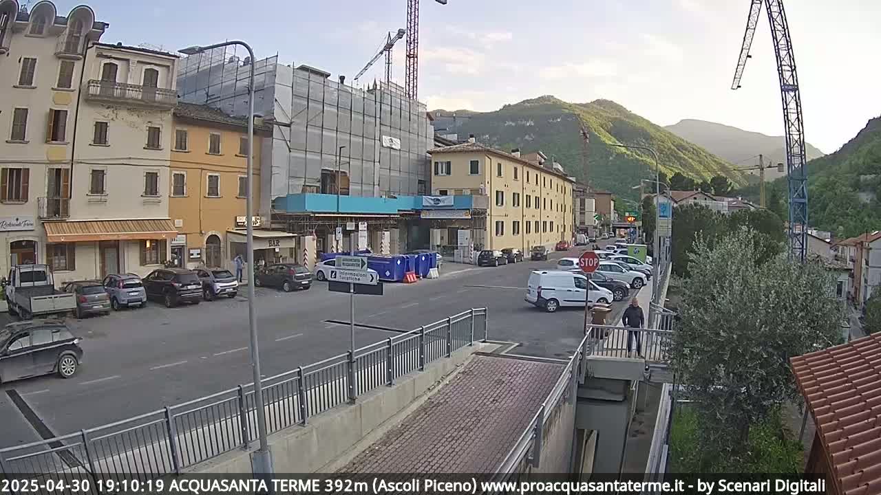 A partly cloudy day reveals a town square with parked cars, several buildings, one under construction, and a pedestrian crossing a footbridge.