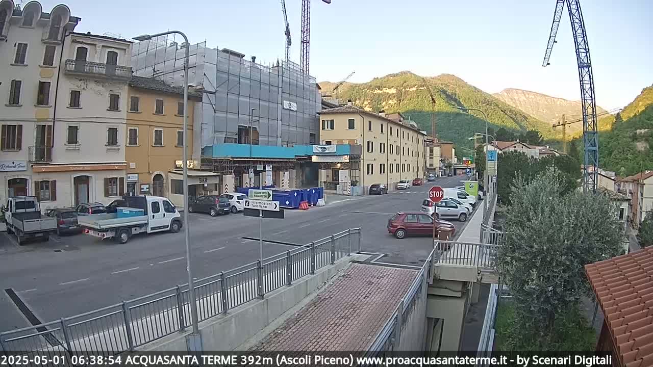 A street scene shows several parked cars and a few buildings under construction, with mountains in the background under a clear sky.