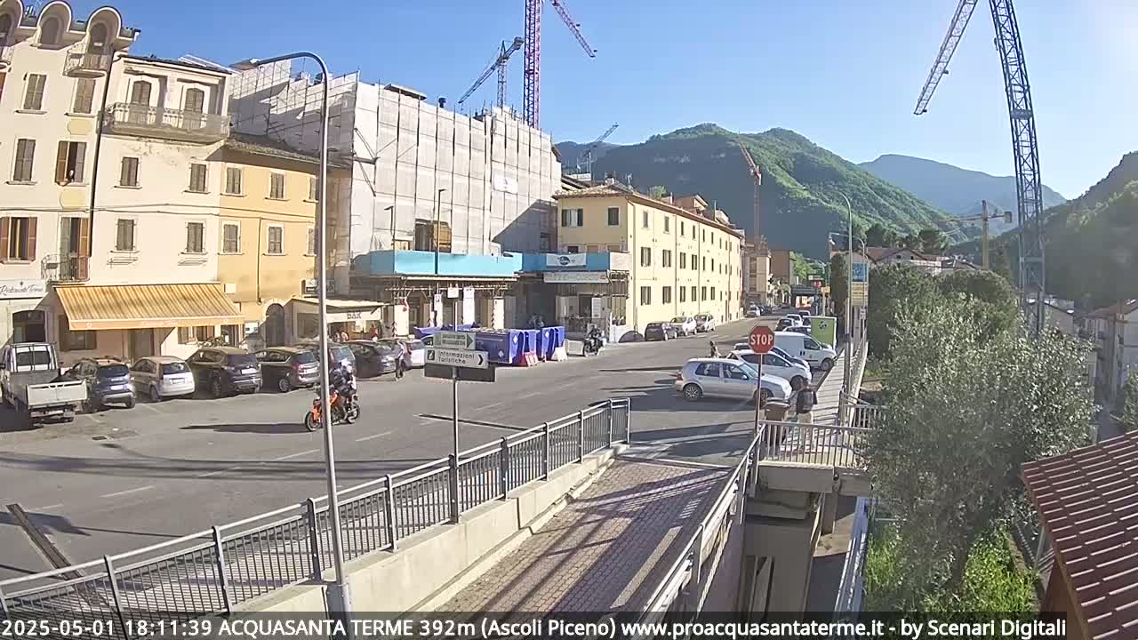 A sunny day reveals a street scene in a town nestled against mountains, with construction underway on a large building, cars parked along the side of the road, and a motorcycle driving down the street.