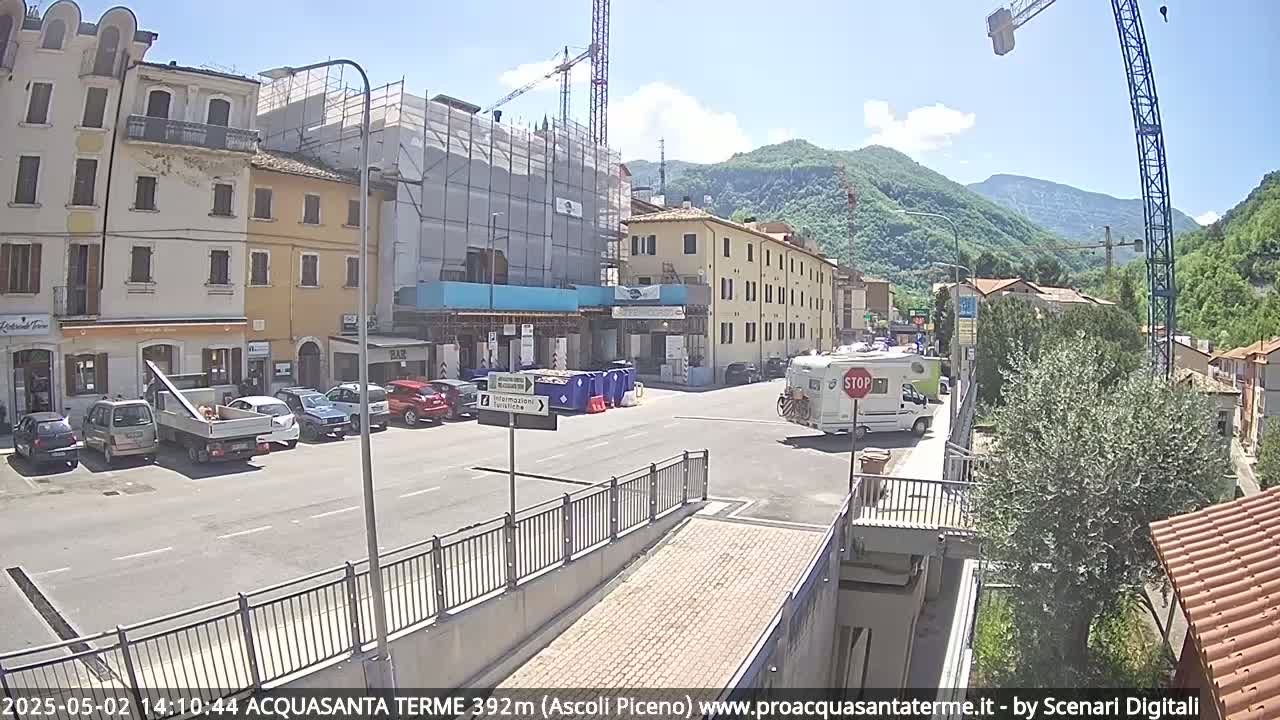 A sunny day reveals a town square with parked cars, a construction site, and a camper van at an intersection, all nestled against a mountainous backdrop.