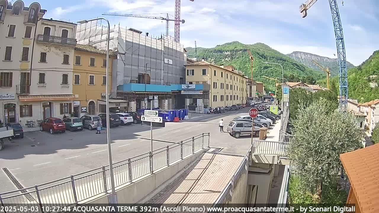 A street scene in a town nestled in a valley under a mostly sunny sky, shows buildings, parked cars, and construction activity.