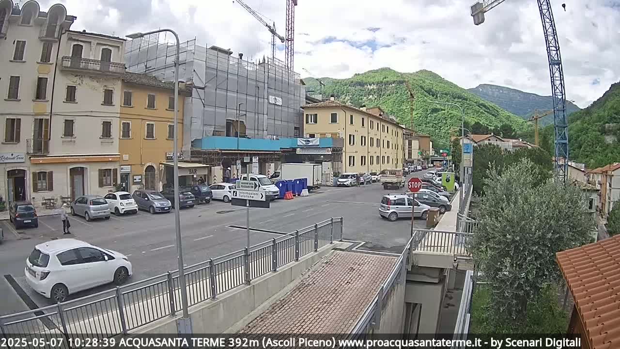 A partly cloudy day reveals a town scene with parked cars, buildings under construction, and a crane, all situated near a pedestrian bridge and hills in the background.