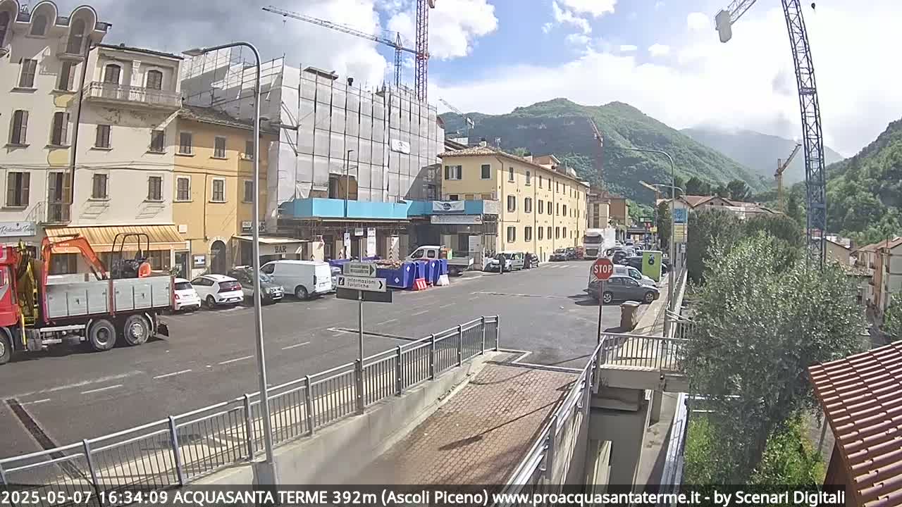 A sunny day reveals a town square with several buildings, one under construction, vehicles parked along the street, and mountains in the background.