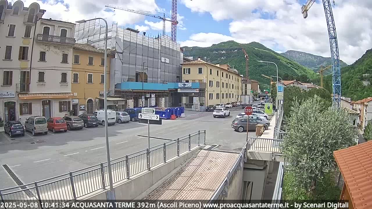 A partly cloudy day reveals a town square with parked cars, buildings under construction, and a crane against a backdrop of green hills.