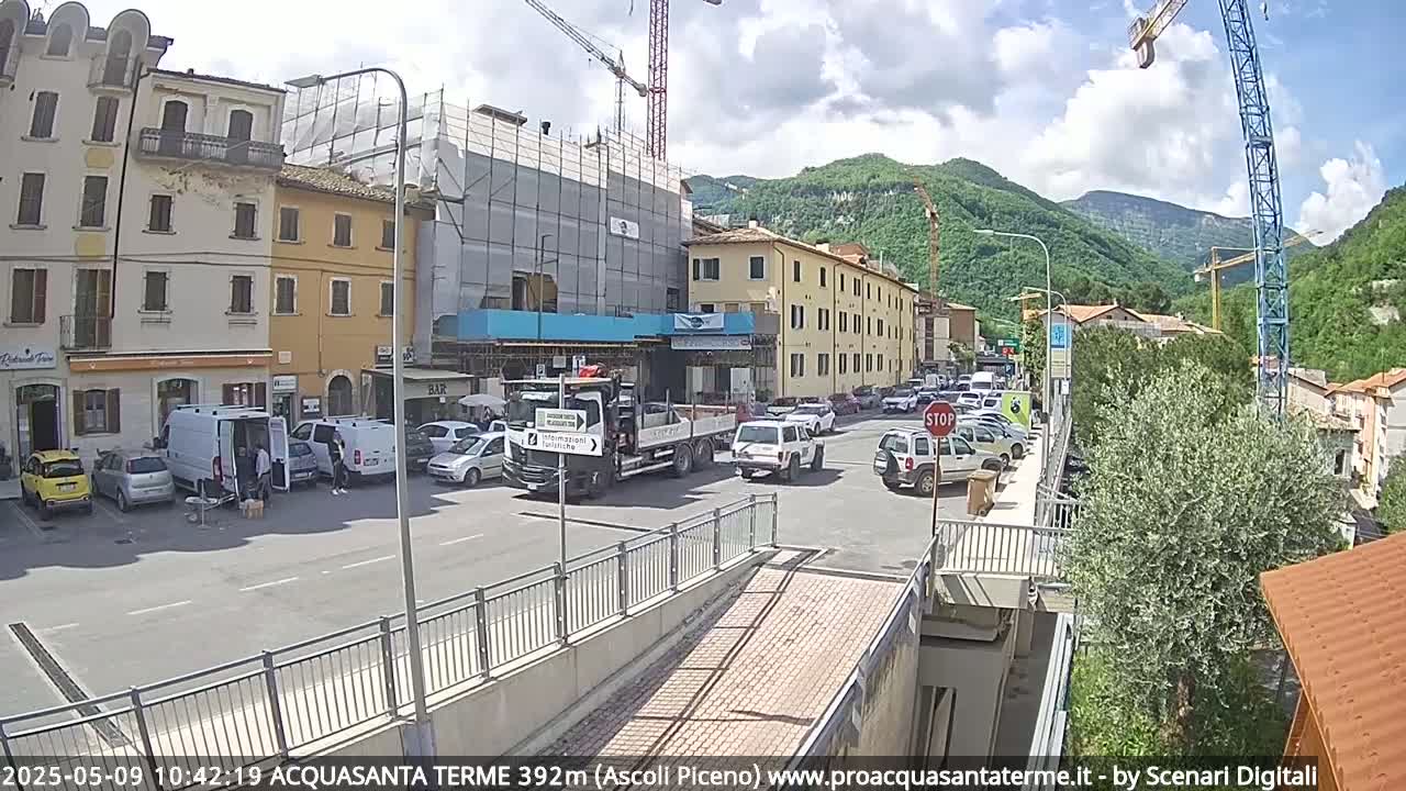 A sunny day reveals a town square with parked cars, a construction site, and a large truck, all situated near mountains under a partly cloudy sky.