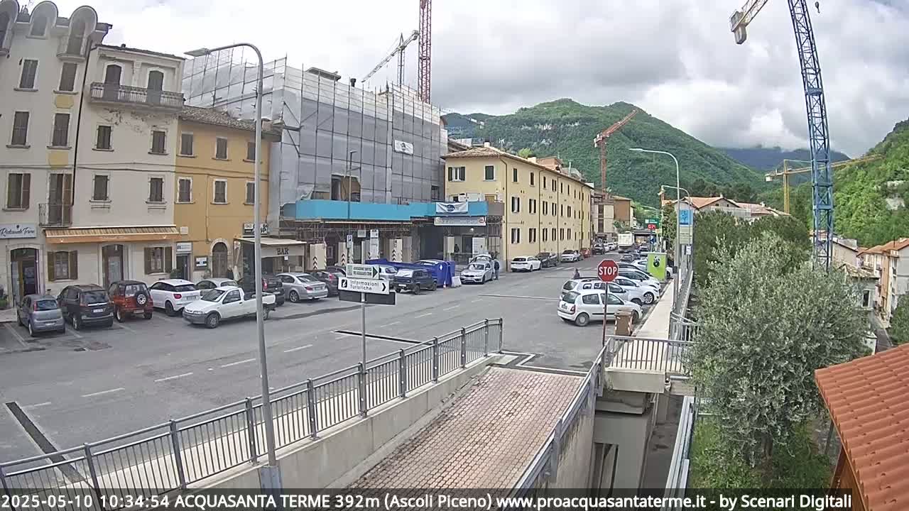 A partly cloudy day reveals a street scene in a town nestled at the base of mountains, with parked cars, buildings under construction, and cranes visible in the background.