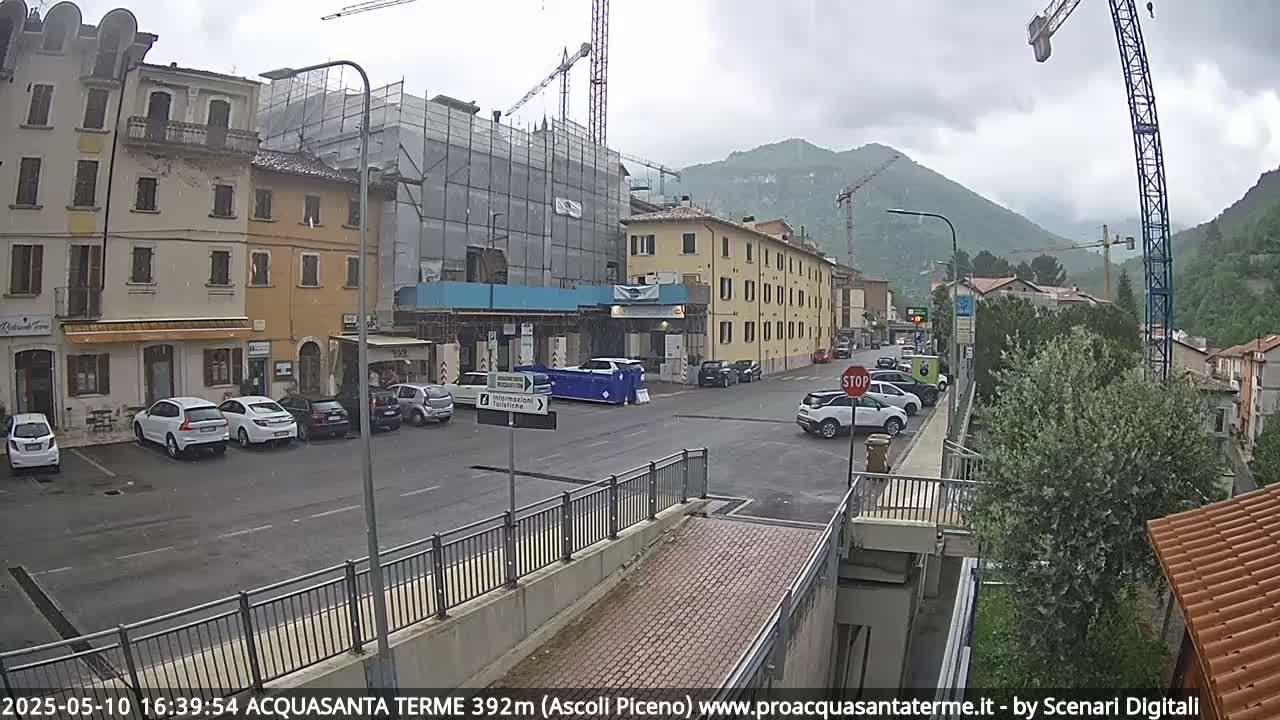A town square with parked cars, buildings under construction, and mountains in the background under an overcast sky.