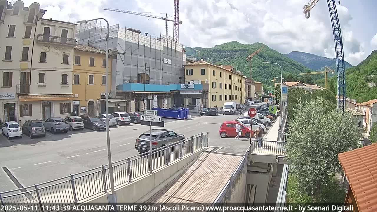 A sunny town square with parked cars, buildings under construction, and a pedestrian bridge, nestled in a valley surrounded by mountains.