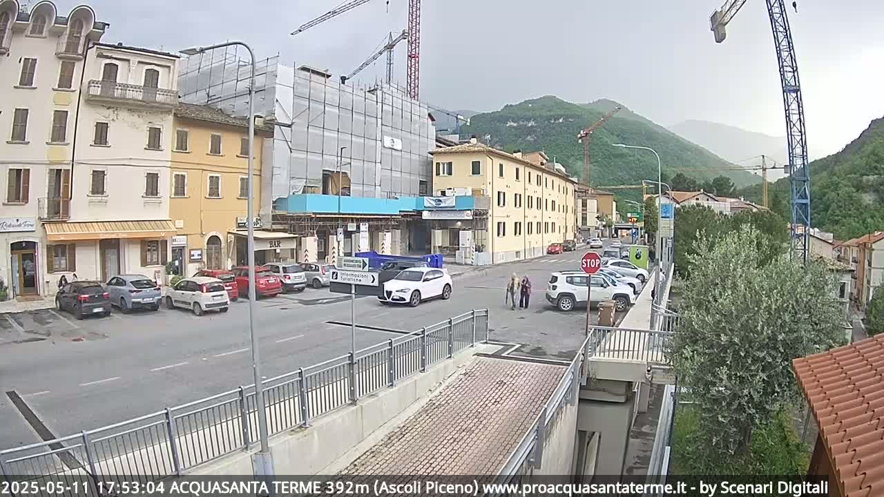 A street scene on an overcast day shows several cars parked and driving near buildings under construction, with mountains visible in the background.