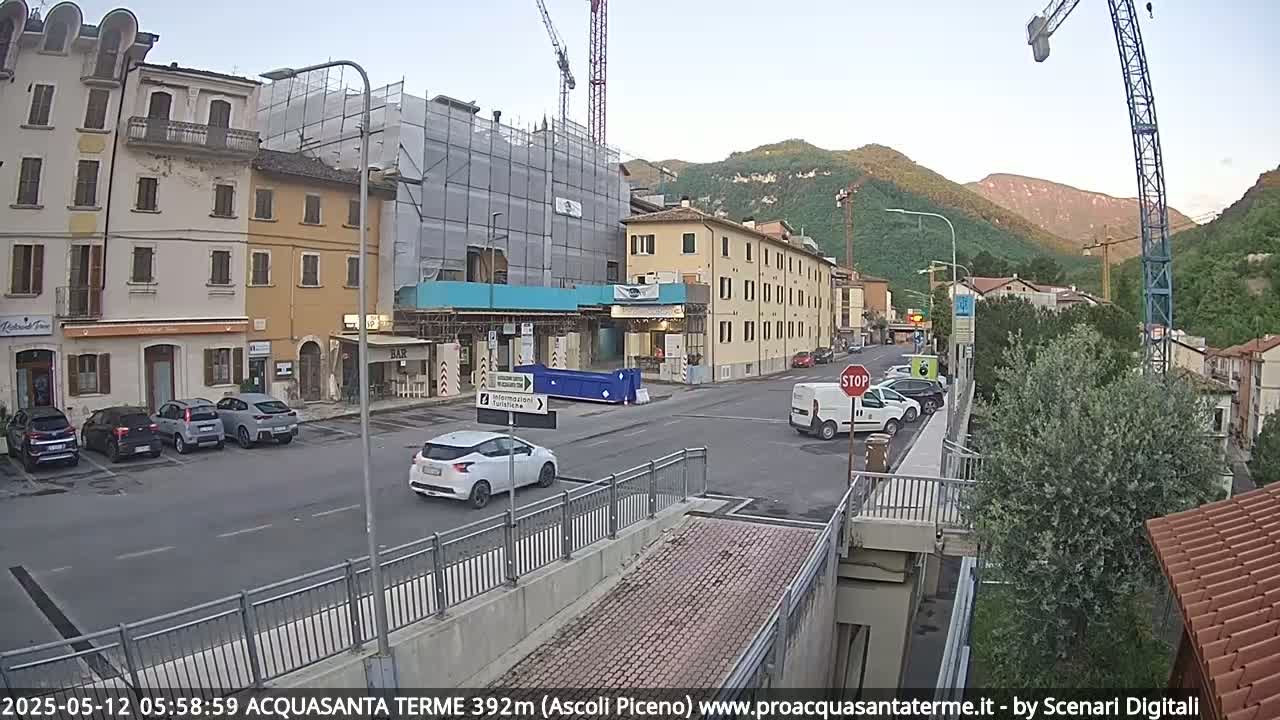A partly cloudy morning scene shows a street with parked and moving cars, several buildings including one under construction, and a crane in the background against a backdrop of hills.