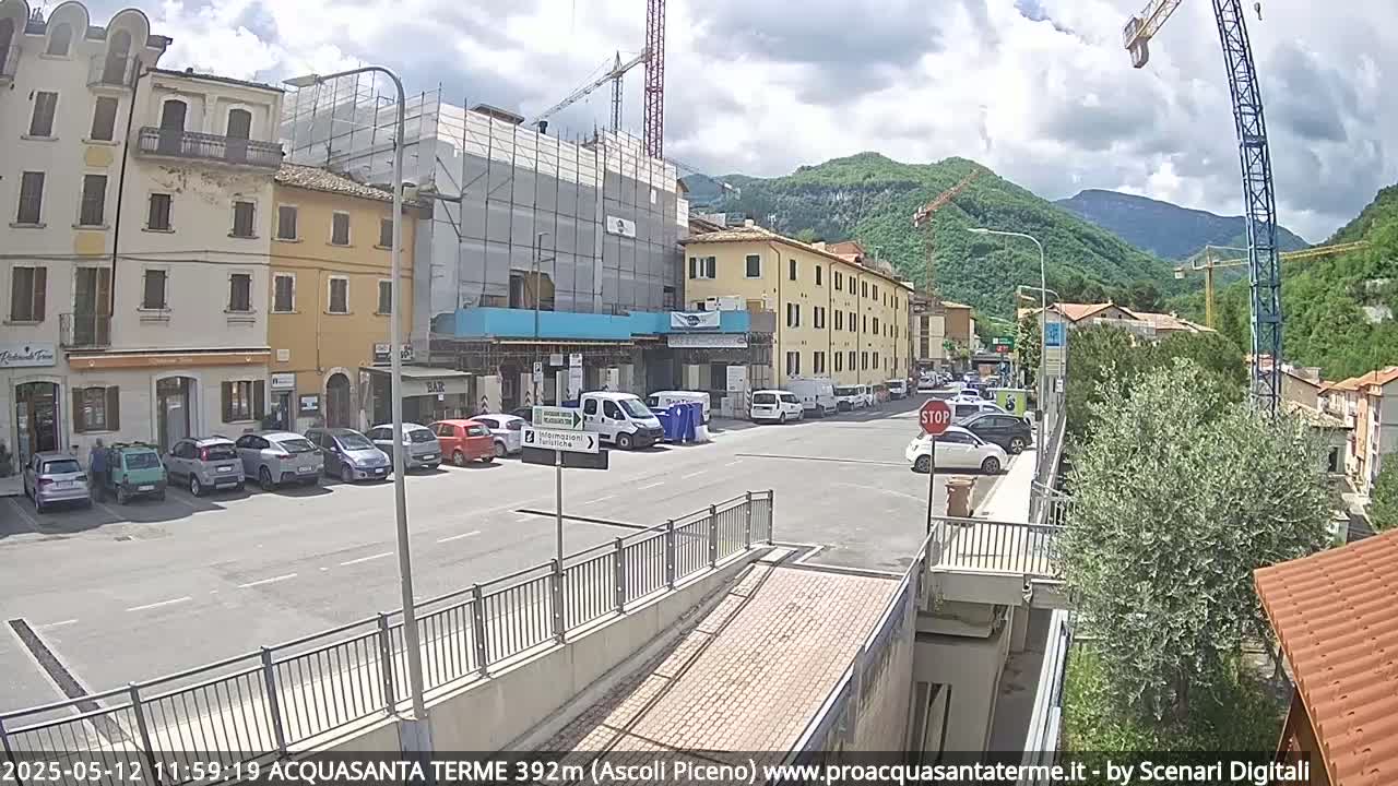 A partly cloudy day reveals a town square with parked cars, buildings under construction, and mountains in the background.