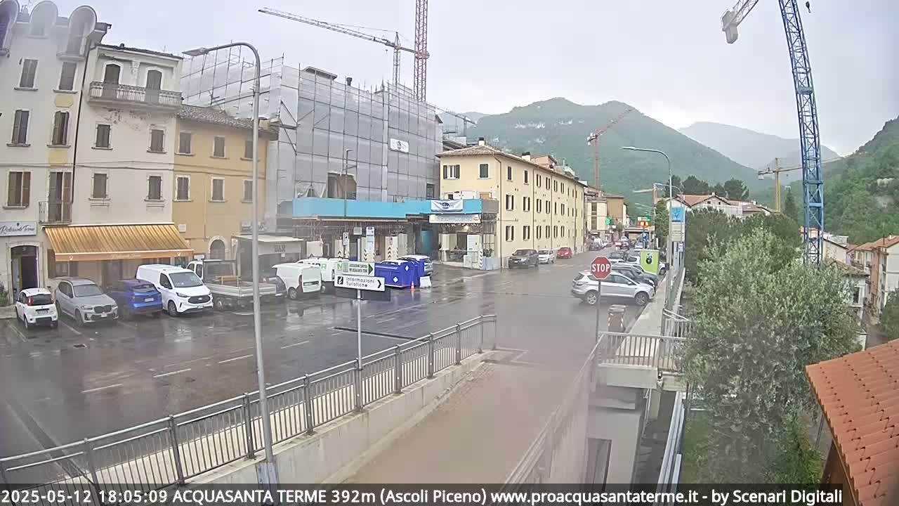 A wet street scene shows several cars parked in front of buildings under construction, with mountains in the background on an overcast day.