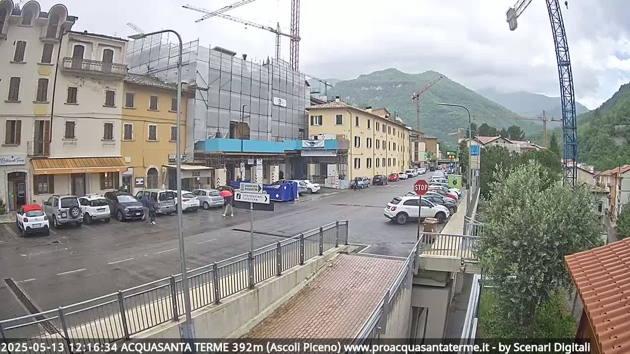 A rainy town square with parked cars, buildings under construction, and mountains in the background.