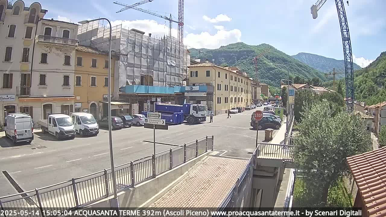 A sunny day reveals a town square with parked vehicles, construction equipment, and buildings, one of which is under construction,  surrounded by hills.