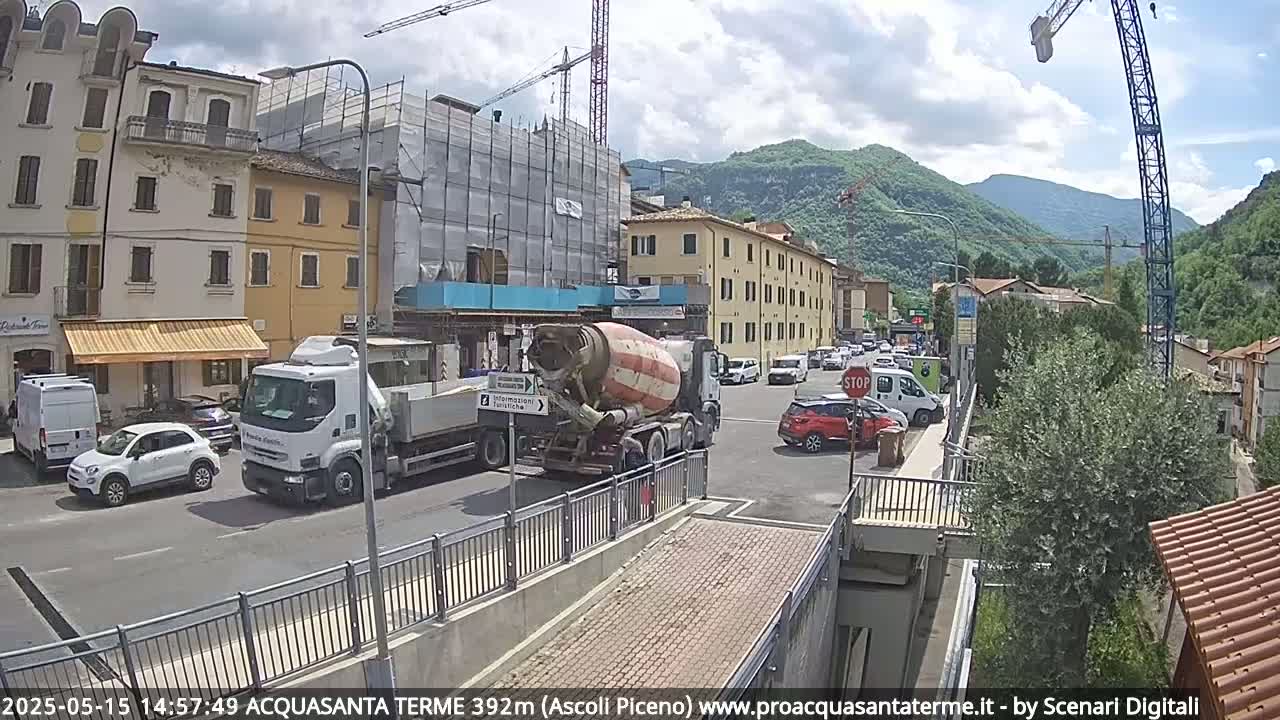 A partly cloudy day shows a street scene with several vehicles, including a cement mixer, near buildings under construction and others along a hillside in a mountainous area.