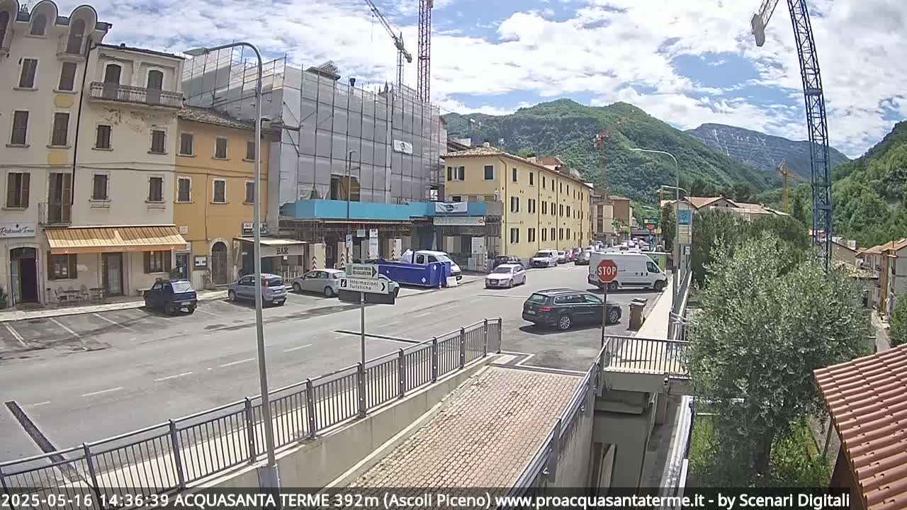 A partly sunny day reveals a town square with several parked cars and buildings, one under construction, nestled in a valley surrounded by mountains.
