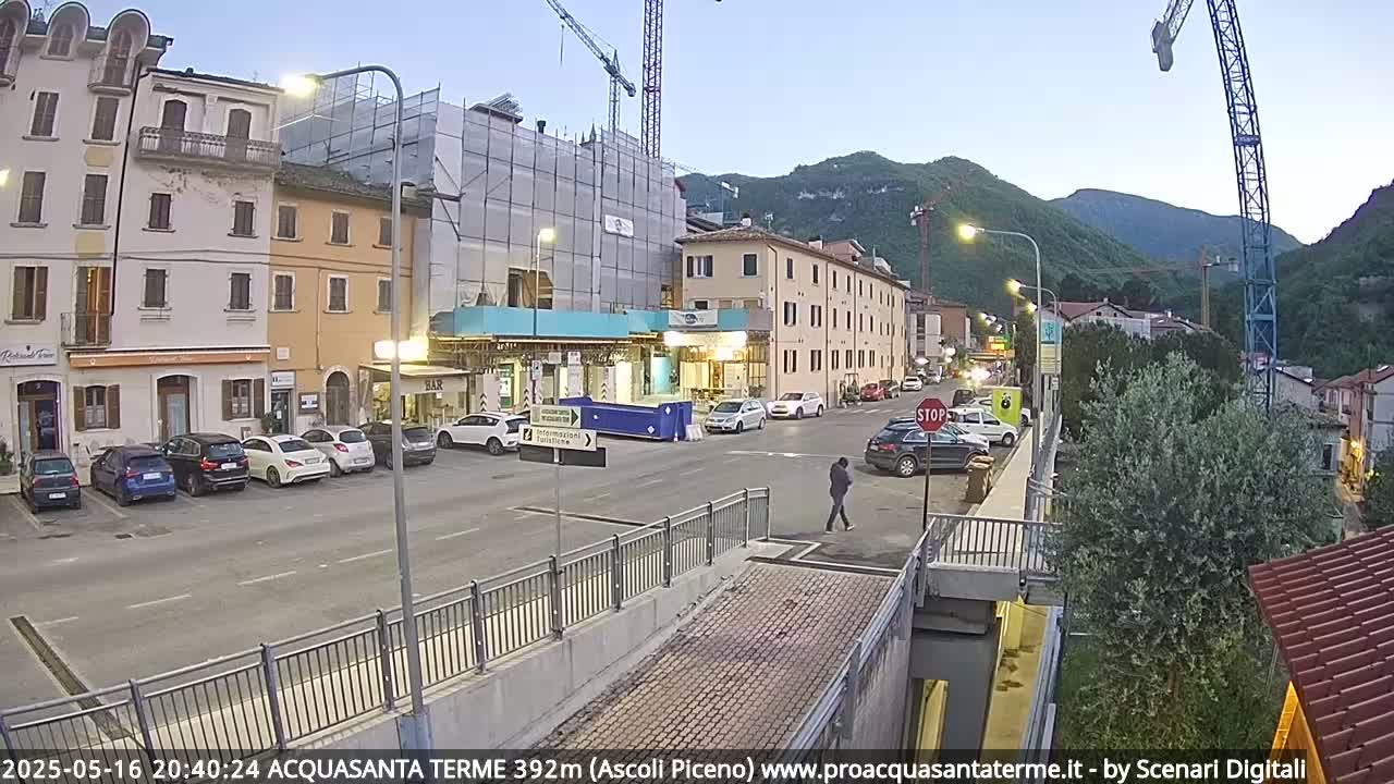 A street scene at dusk shows several cars parked along the sides of buildings under construction and undergoing renovation, with hills and mountains in the background.