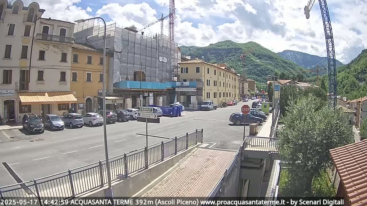 A partly sunny day shows a town square with parked cars, buildings under construction, and a large crane, all situated near a mountain range.