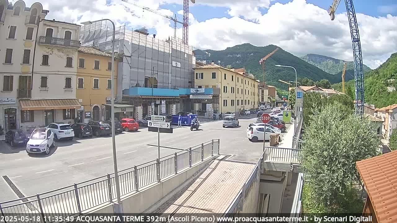 A sunny day in a town with buildings under construction, parked cars, and some pedestrians and vehicles in motion, all set against a backdrop of mountains.