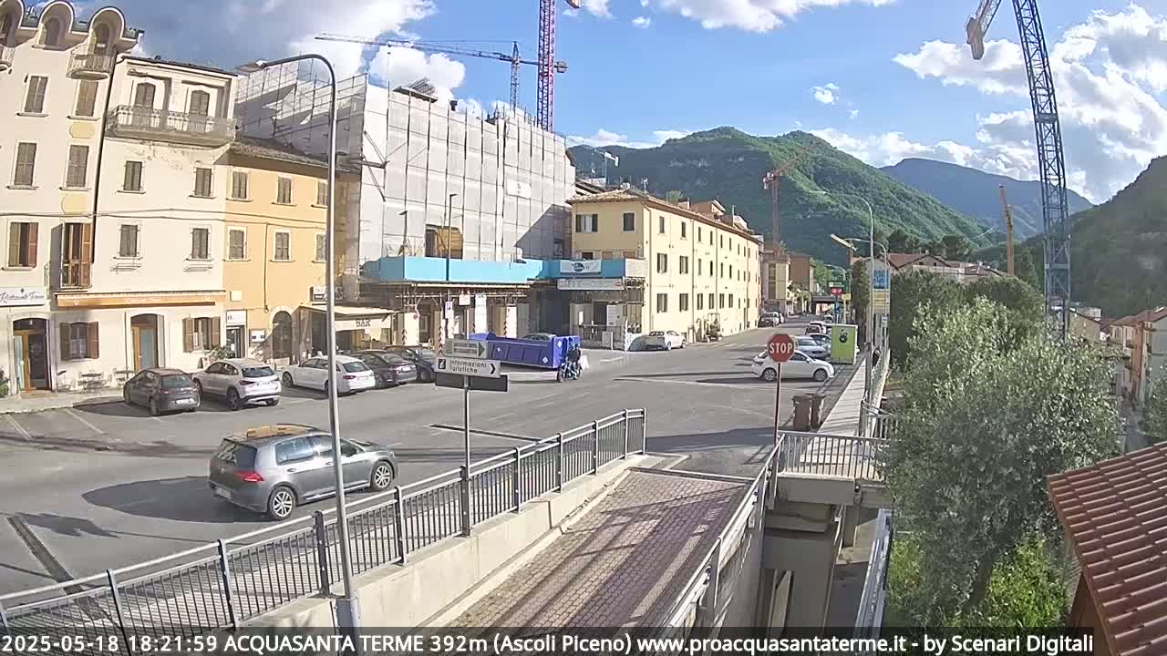 A partly sunny day reveals a town square with parked cars, buildings under construction, and mountains in the background.