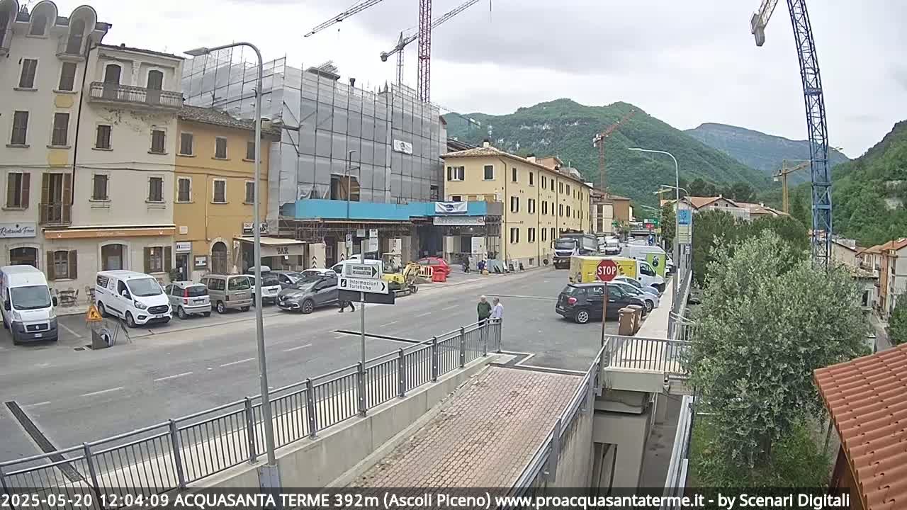 A street scene shows several cars parked along the side of the road in front of buildings under construction, with mountains visible in the background on a partly cloudy day.
