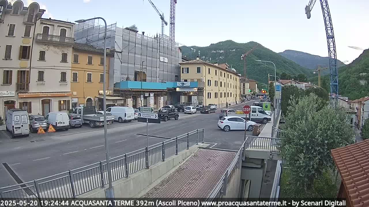 A street scene in a town nestled against mountains shows several cars parked and driving, with buildings under construction and a crane visible in the background under a partly cloudy sky.