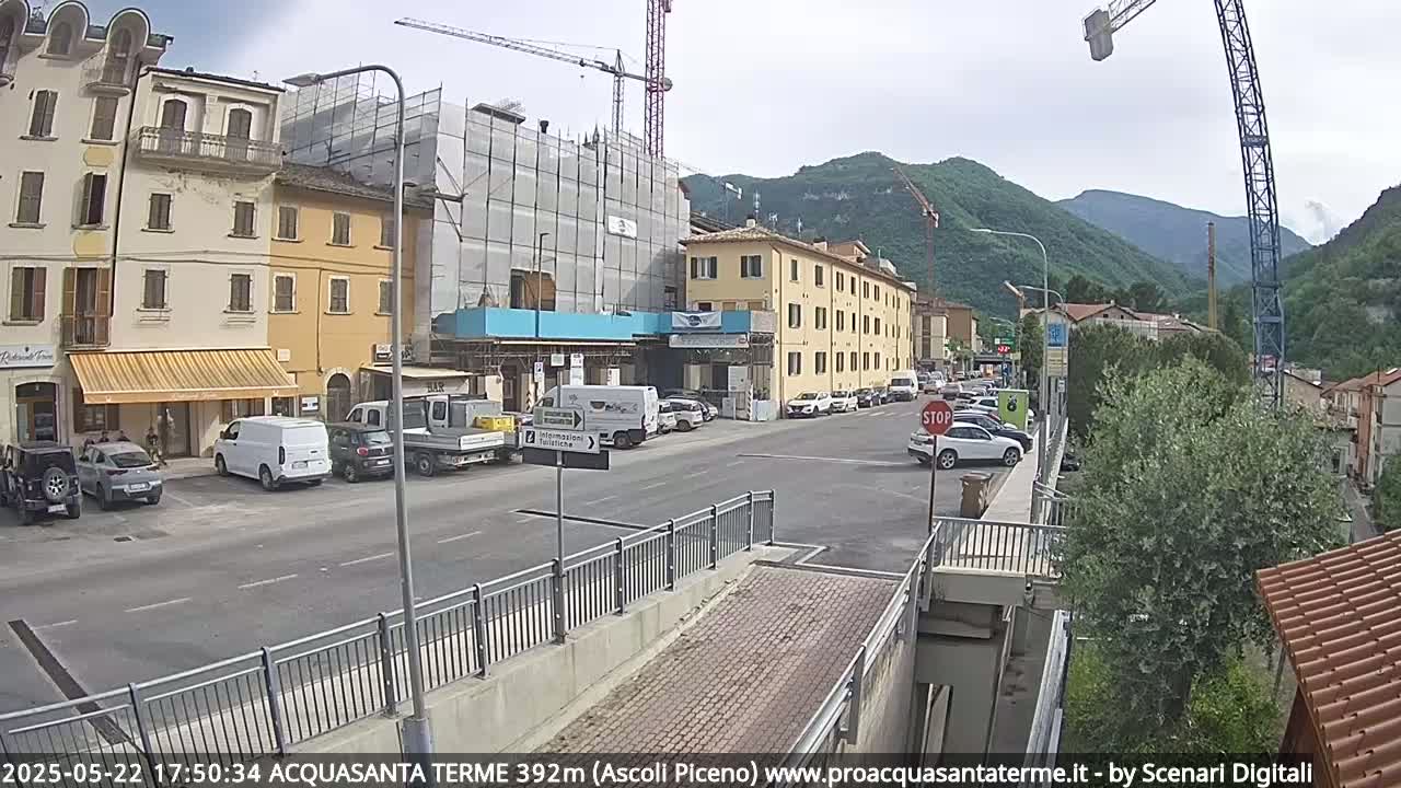 A street scene on a partly cloudy day shows several parked cars, buildings under construction, and a few pedestrians near mountains in the distance.