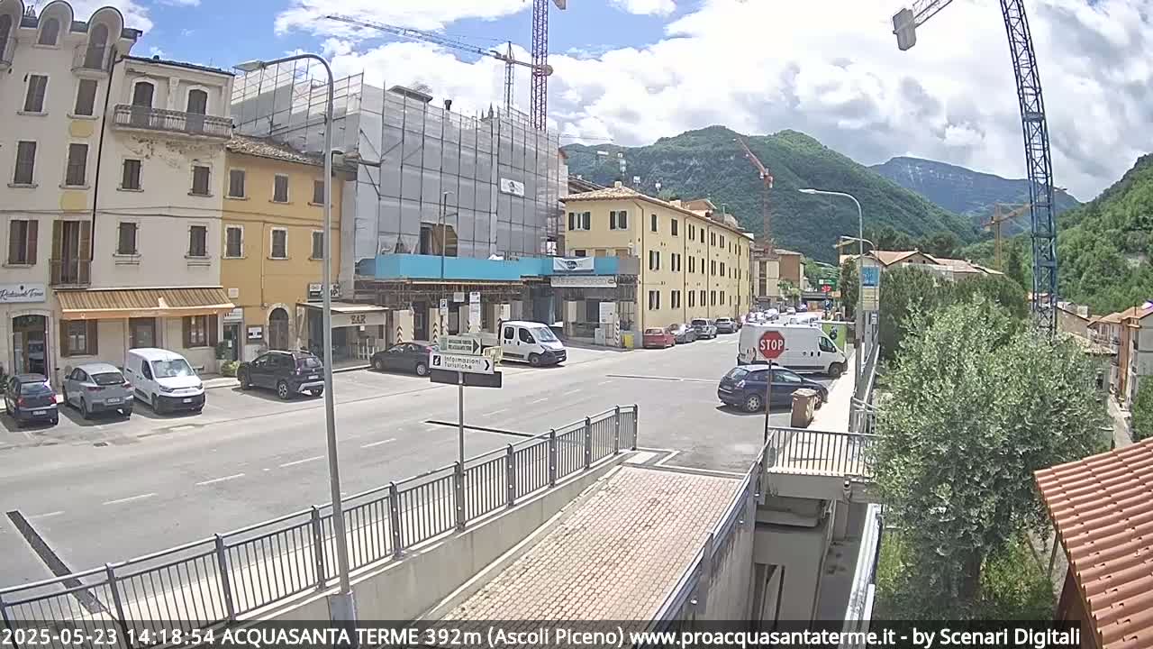 A partly cloudy day reveals a street scene in a town nestled in a valley, with several buildings, parked cars, and construction underway, including a crane.