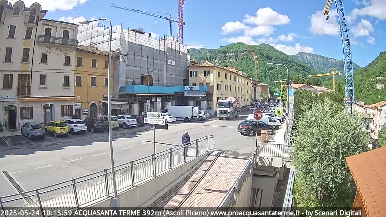 A sunny day reveals a town square with parked cars, construction scaffolding on a building, and cranes against a backdrop of green hills.
