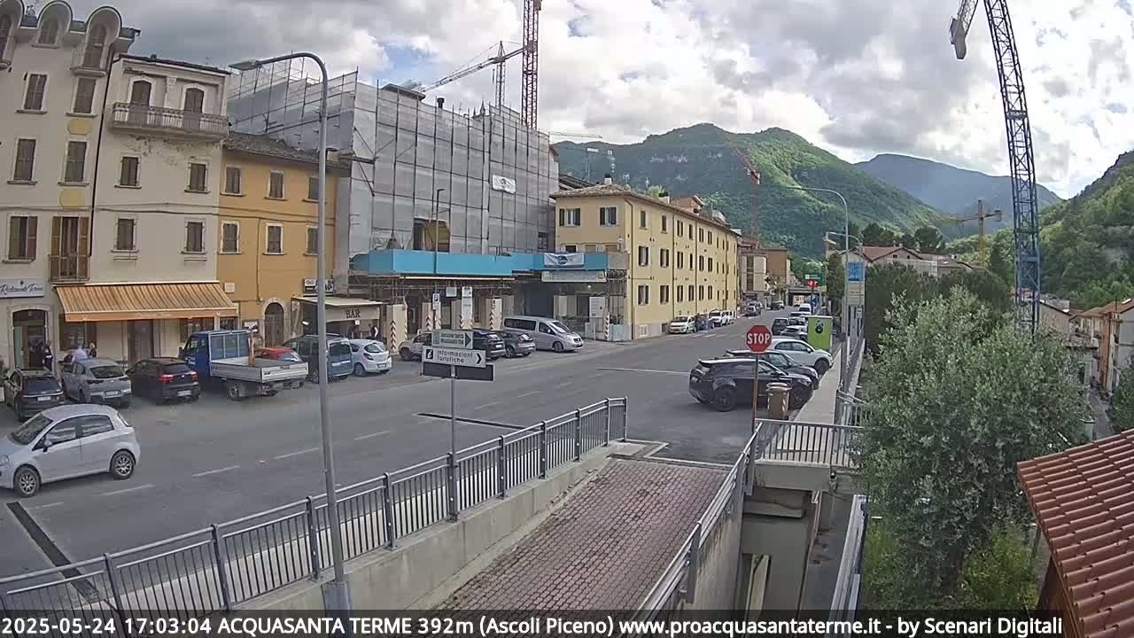 A partly cloudy day reveals a town street scene with parked cars, buildings under construction, and mountains in the background.