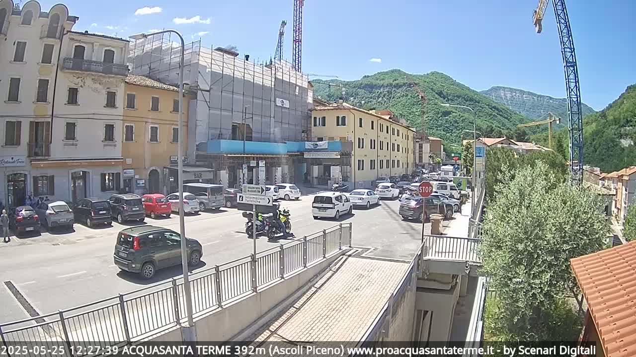 A sunny day shows a town square with parked cars, motorcycles, and some traffic, with construction scaffolding on a building and a large crane in the background, all surrounded by hills.