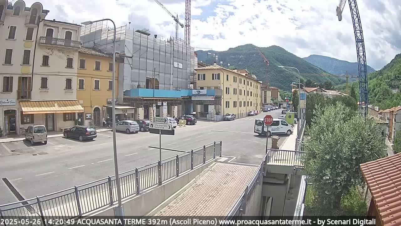A partly cloudy day reveals a town square with parked cars and buildings, one of which is under construction, set against a backdrop of mountains.