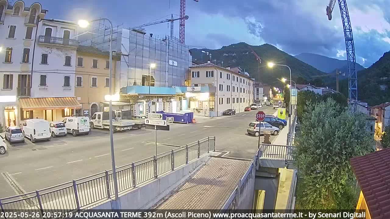 A town square at dusk, with parked cars and buildings, including one under construction, situated against a backdrop of mountains under a partly cloudy sky.