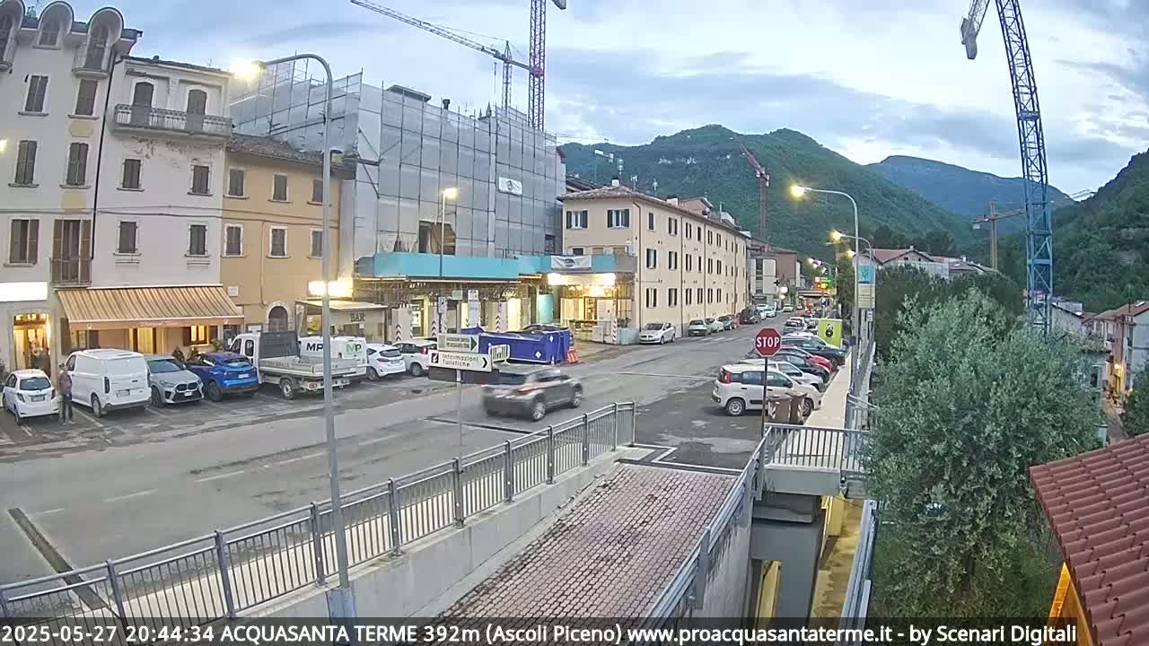 Cars are parked along the side of a street in a town nestled in a valley at dusk, with a building under construction visible in the background.