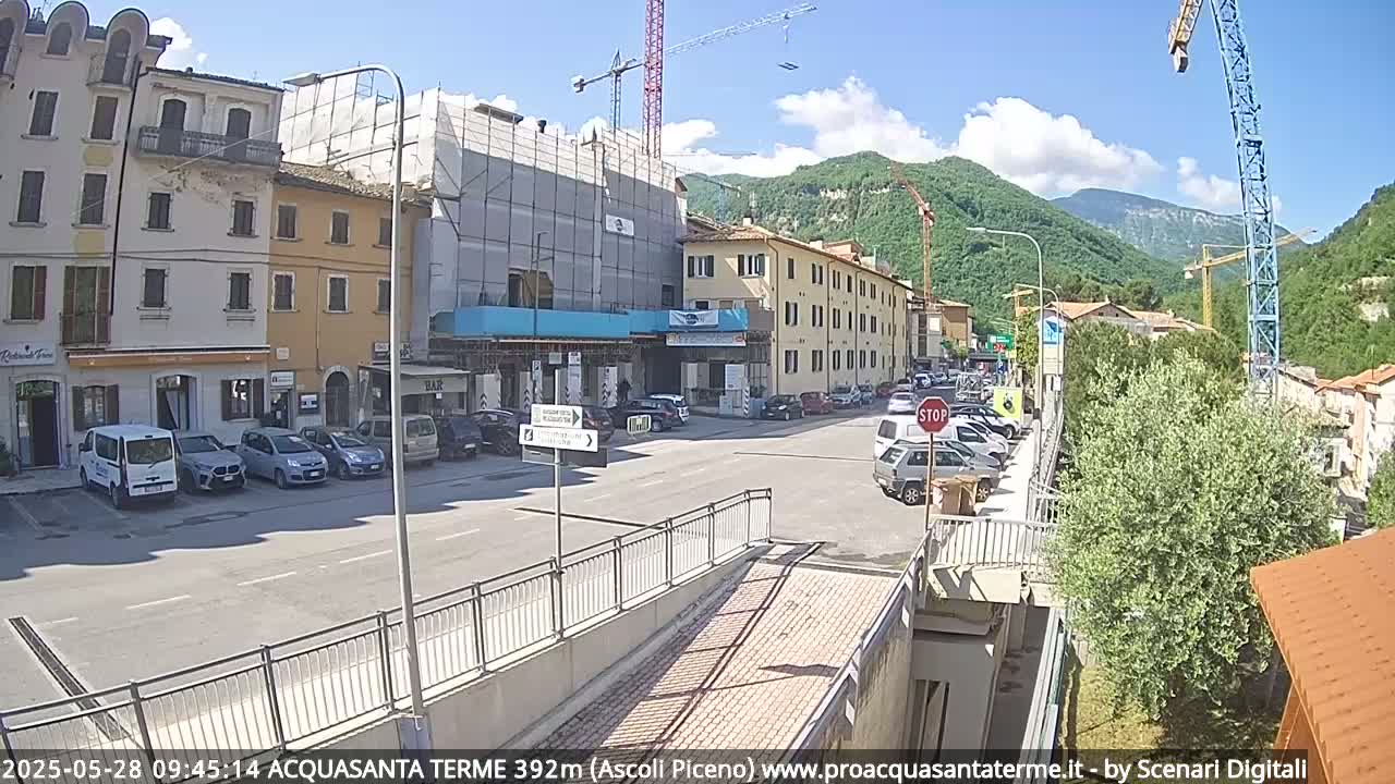 A sunny day in a town with buildings under construction, parked cars, and mountains in the background.