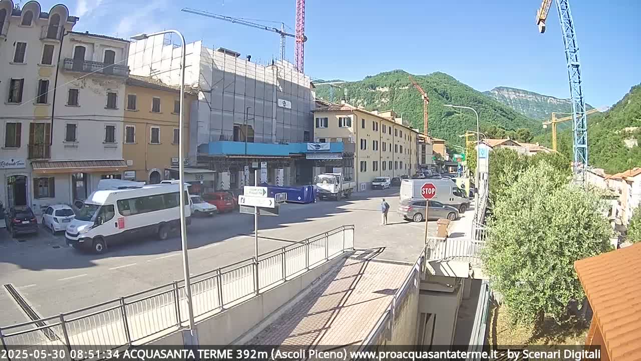 A sunny town square scene shows several buildings, some under construction, vehicles parked along the street, and a person walking near a stop sign, with hills and construction cranes visible in the background.