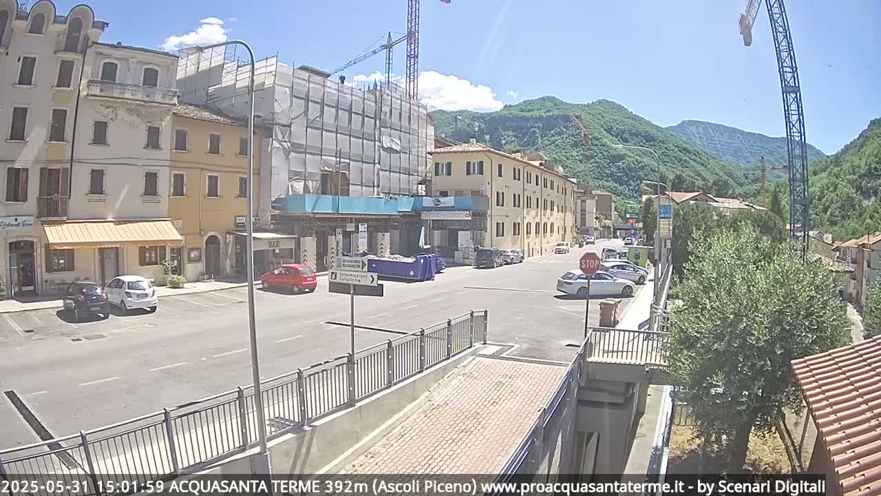 A sunny day reveals a town square with parked cars, buildings under construction, and mountains in the background.