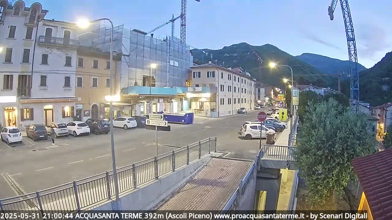 A street scene at twilight shows several parked cars in front of buildings under construction, with hills in the background.