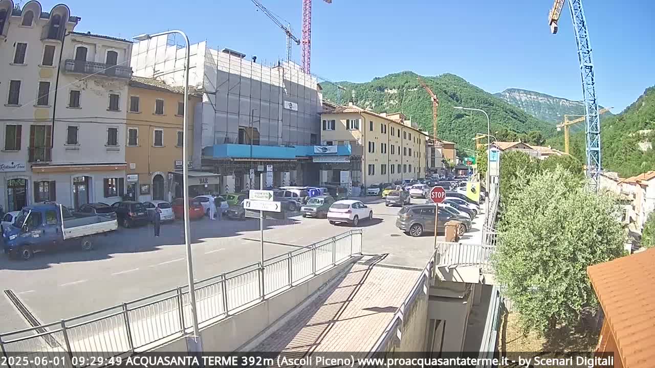 A sunny day reveals a town square with parked cars, buildings under construction, and construction cranes visible against a backdrop of green hills.